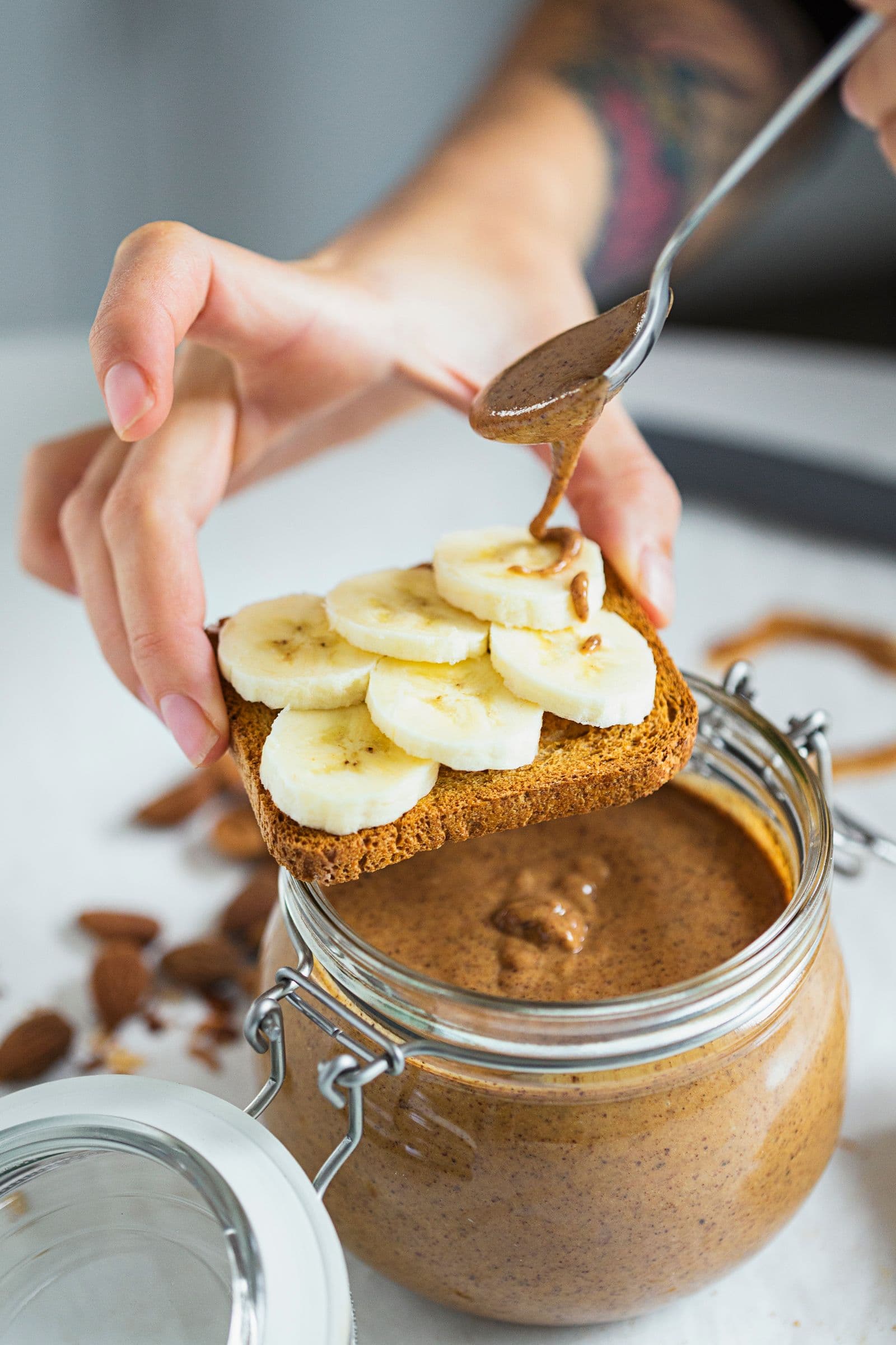Hand drizzling almond butter on toast with sliced bananas over an open jar.