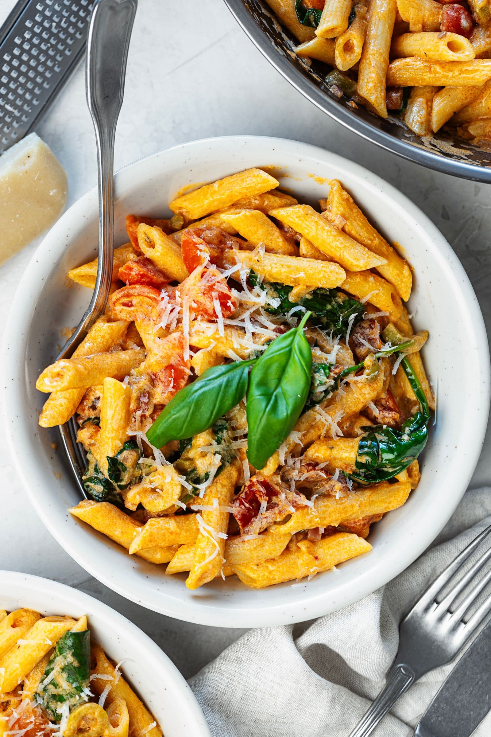 Top-down view of a bowl of creamy tomato and spinach penne next to the serving pan, garnished with fresh basil
