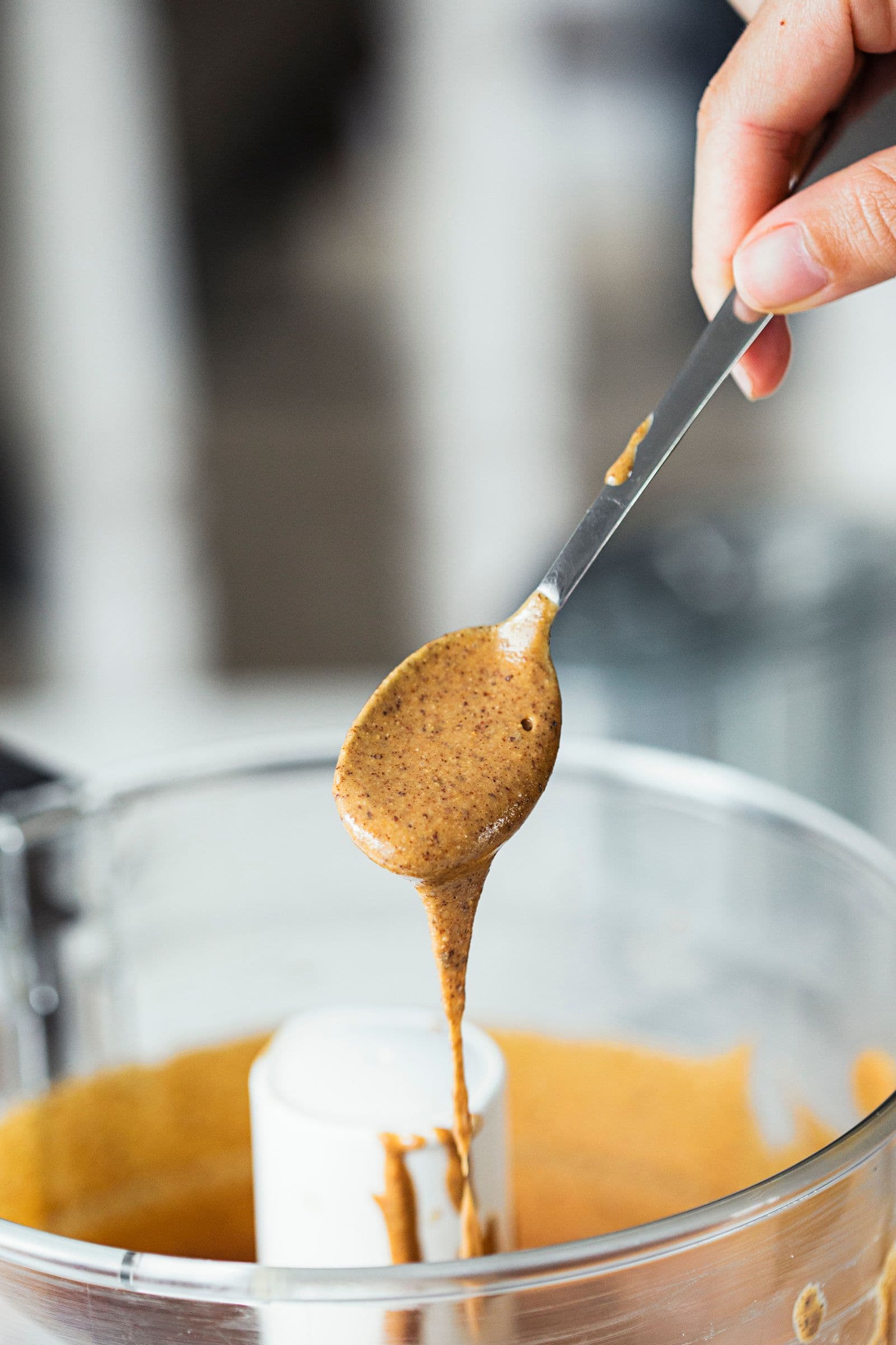 Spoon drizzling almond butter into the food processor jar.