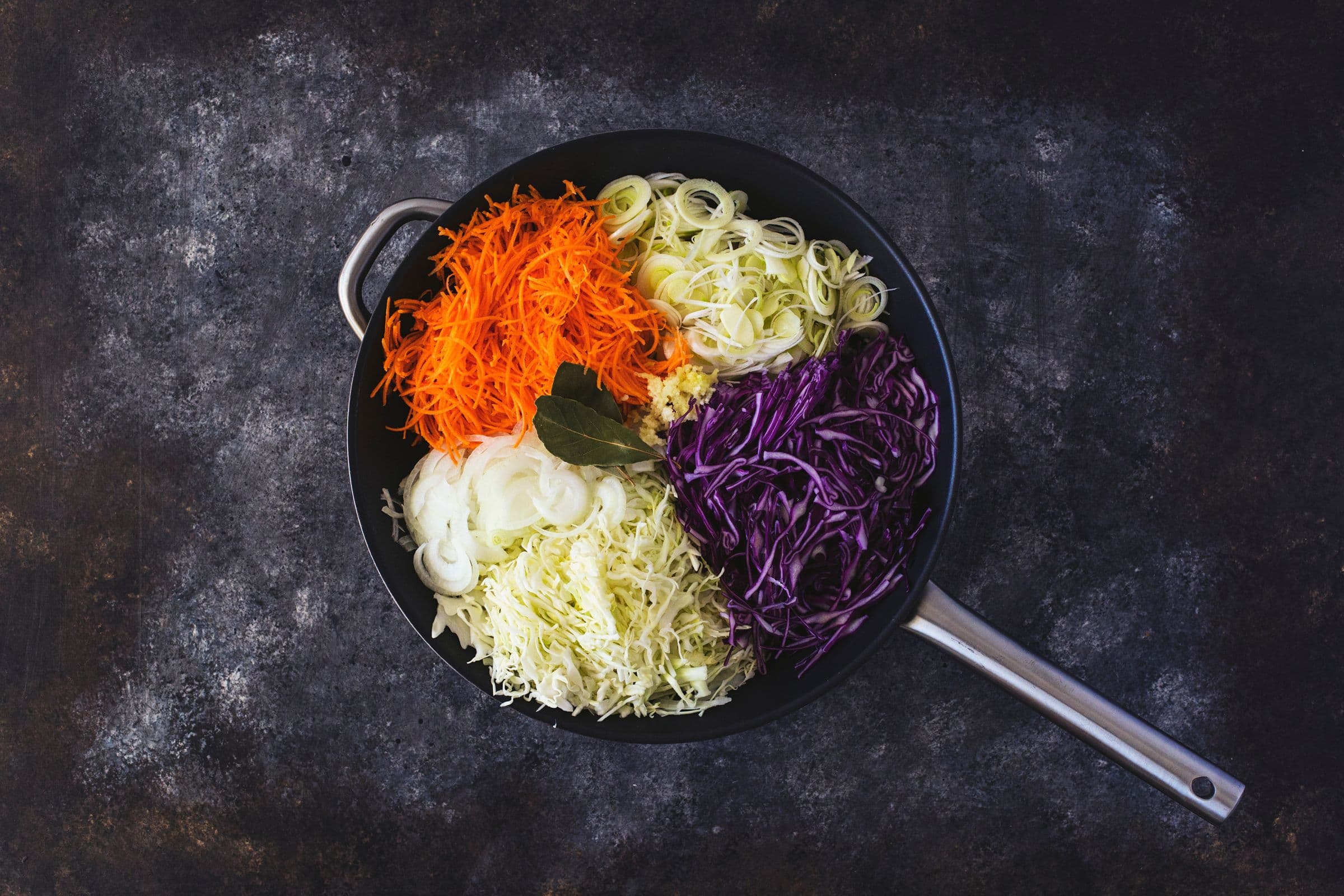 Black frying pan filled with grated carrots, thinly sliced onions, leeks, and cabbage, ready to cook.